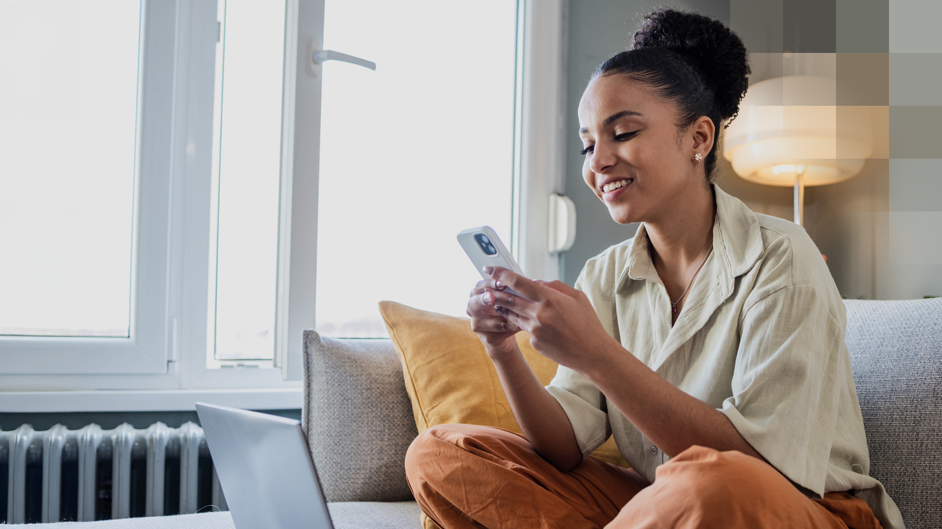 Eine junge Frau sitzt auf dem Sofa, hält ihr Smartphone und hat ein Laptop vor sich, und informiert sich über die Spitaltaggeldversicherung.