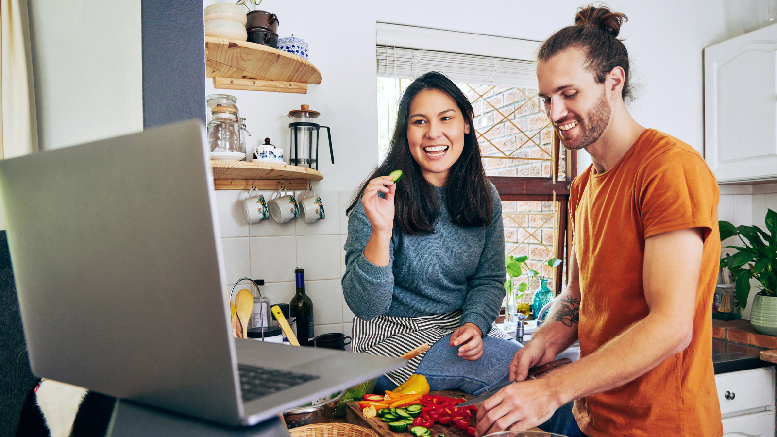 Un homme et une femme se tiennent dans la cuisine, sourient en cuisinant et regardent sur leur ordinateur comment perdre la graisse abdominale de manière saine.