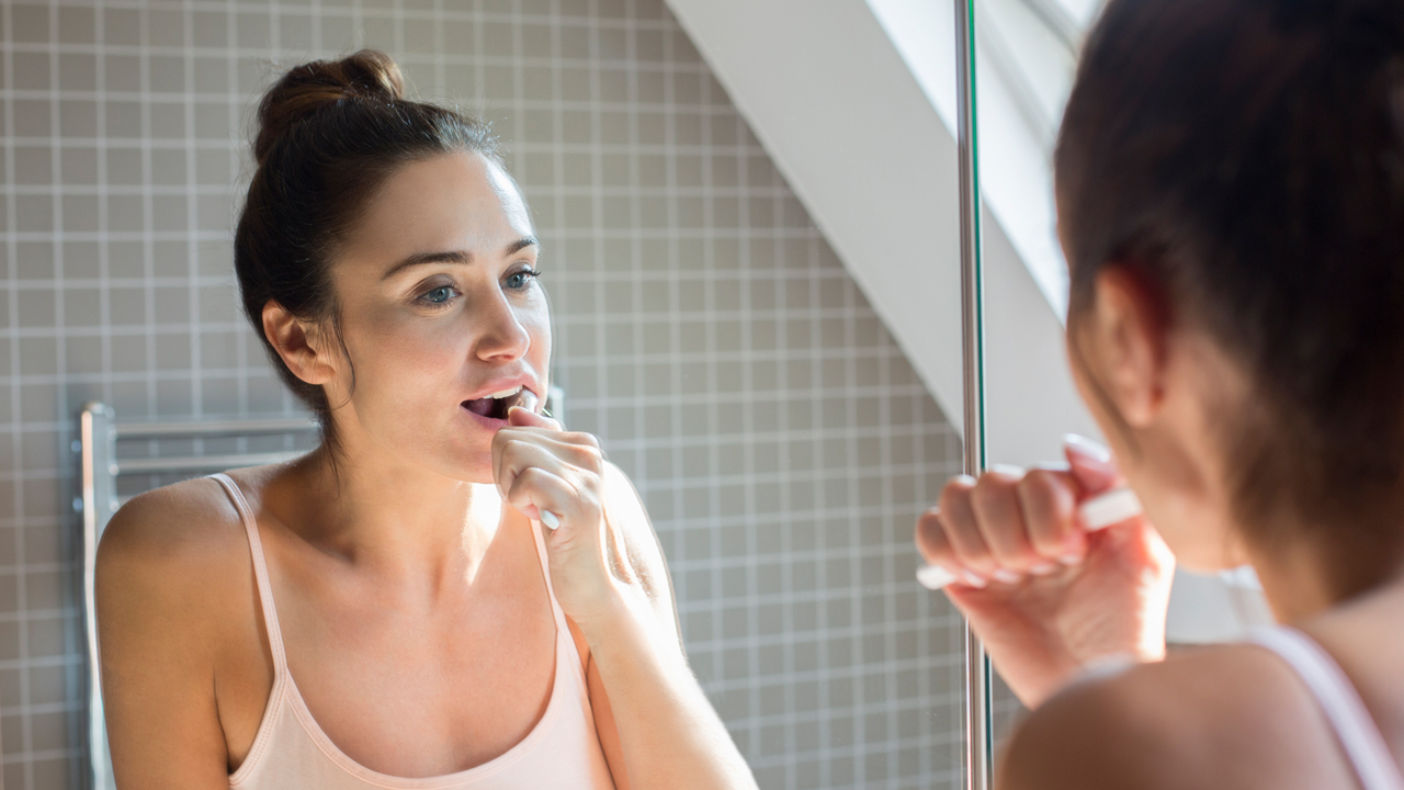 Une femme aux cheveux bruns attachés se brosse les dents devant le miroir de la salle de bains – assurance complémentaire appareil dentaire.