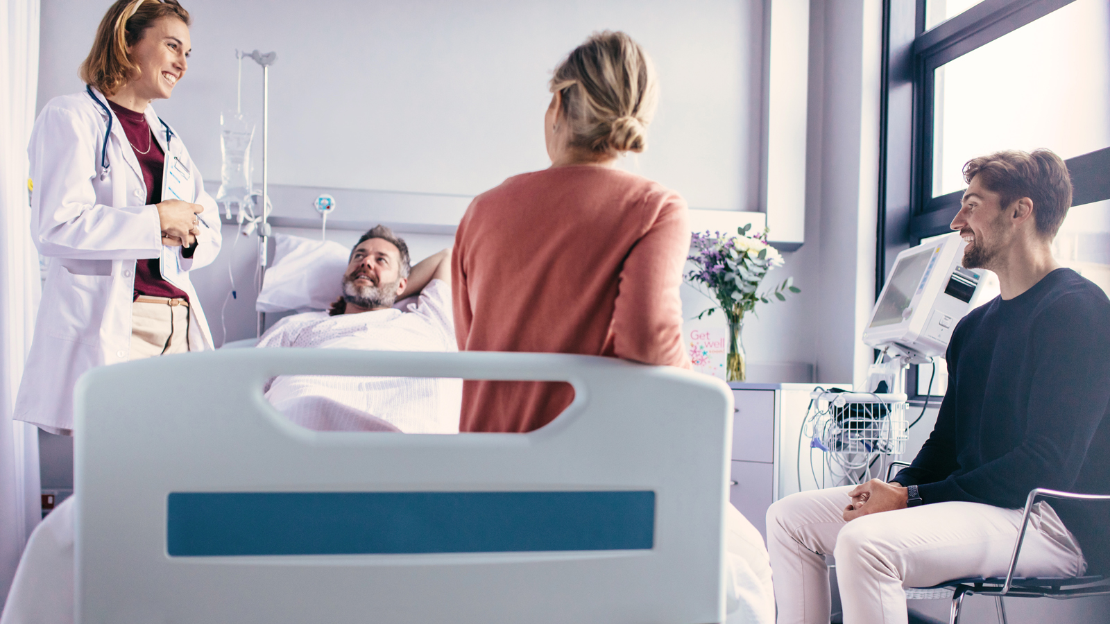 Un homme est allongé dans un lit d’hôpital tandis qu’une médecin souriante discute avec deux visiteurs pour expliquer pourquoi une assurance hospitalière complémentaire est nécessaire et utile.