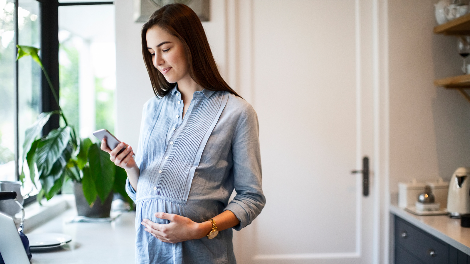 Une femme enceinte se tient dans son appartement et consulte son téléphone pour savoir quelles prestations sont couvertes par l’assurance-maladie pendant la grossesse.