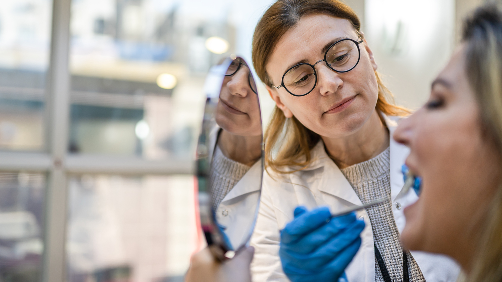 Une dentiste examine la bouche d’une patiente qui tient un miroir et l’informe sur le prix d’un appareil dentaire en Suisse.