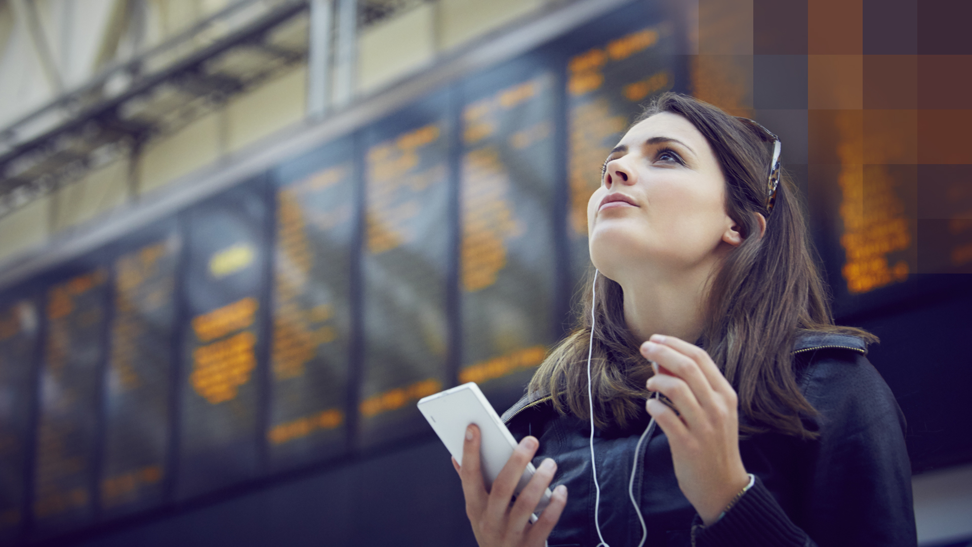 Une jeune femme se tient à l’aéroport devant de nombreux écrans, avec smartphone et écouteurs, et réfléchit à son assurance maladie UE/AELE.